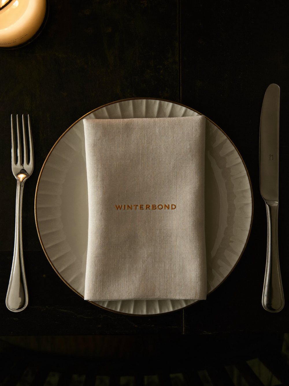 Un cubierto visto desde arriba: un plato con borde dorado y una servilleta de lino doblada, bordada con Winterbond